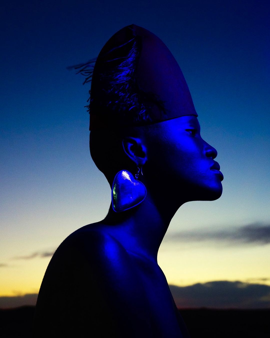 Portrait of a black woman in profile wearing a large heart earring and hat against a blue and yellow gradient sky at dusk.