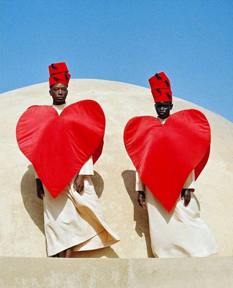 Two models wear oversized red heart costumes and matching headwear against a beige background and blue sky, creating a striking fashion image.
