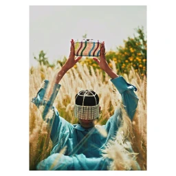 Person in light blue shirt holding a colourful striped clutch above head among tall grass wearing crystal headpiece