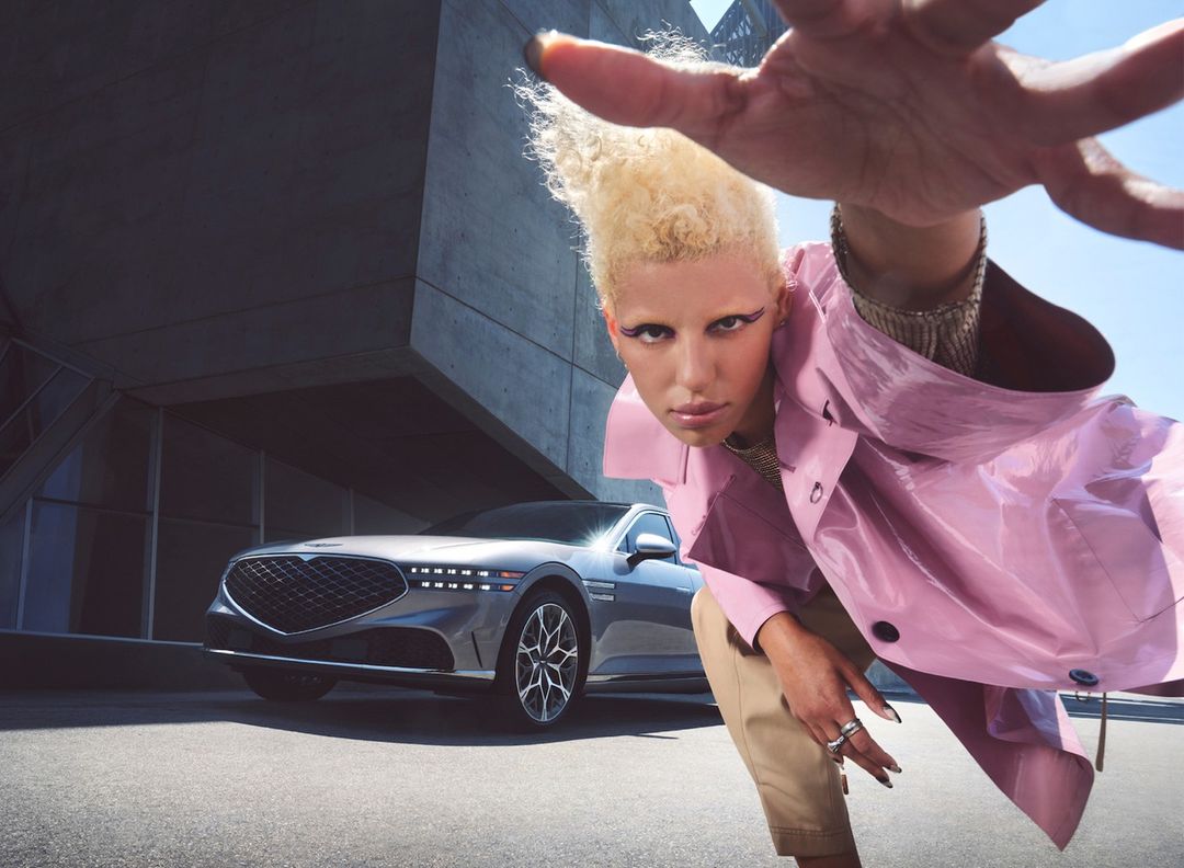 Fashionable person in pink coat and beige pants posing with luxury sedan against a geometric concrete building under clear blue sky.
