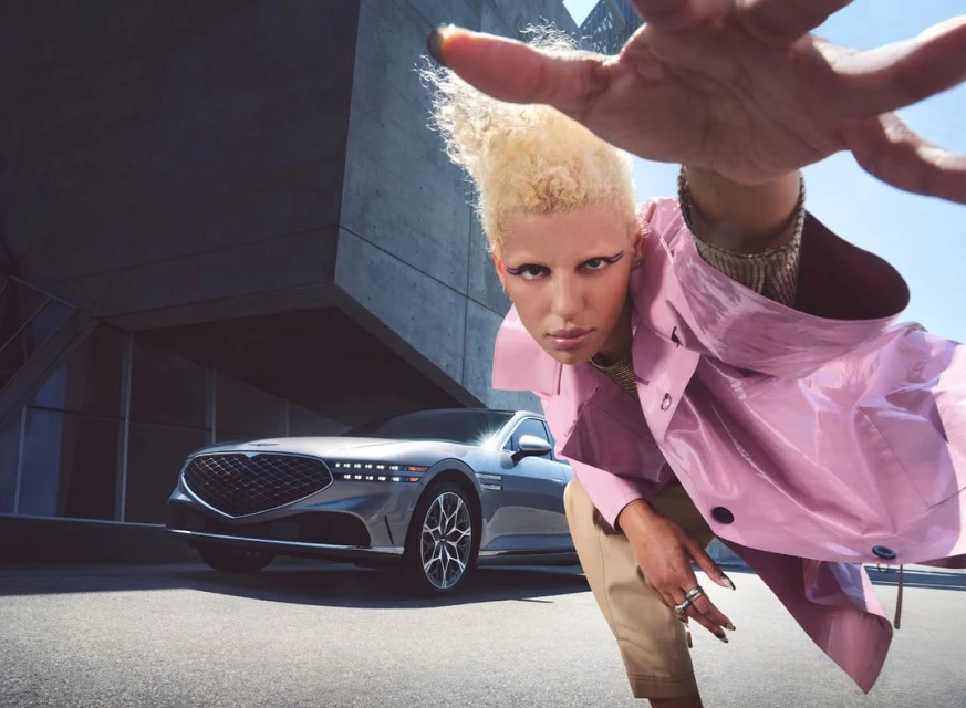 Fashionable person in pink coat and beige pants posing with luxury sedan against a geometric concrete building under clear blue sky.
