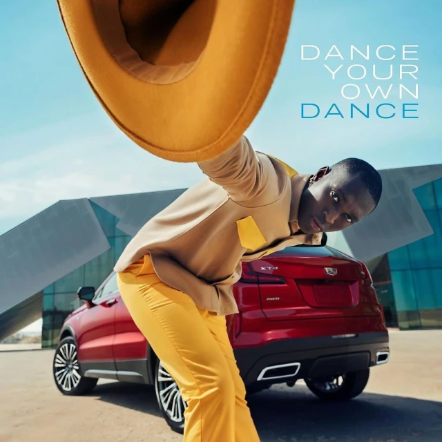 Khaby Lame poses with a tan hat, light brown jacket, and yellow pants, in front of a red Cadillac SUV and modern architecture.