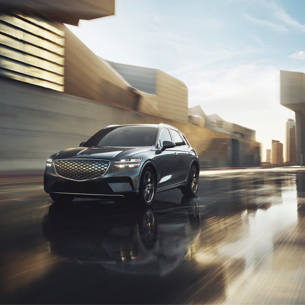 Gray Genesis GV70 SUV driving on a wet city street with blurred motion and modern buildings under a bright sky, automotive photography.