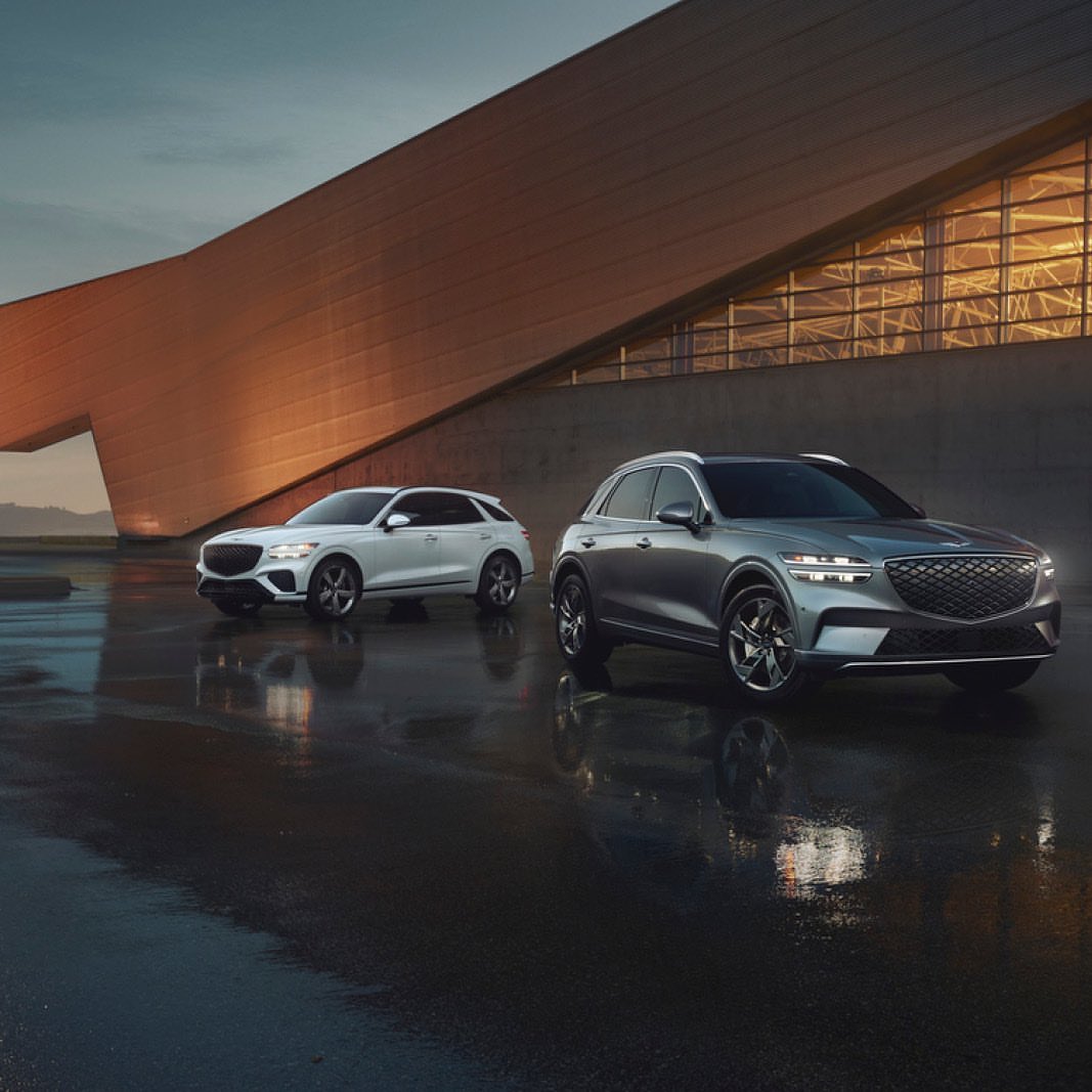 Two Genesis GV70 SUVs parked outside modern building, reflecting on wet ground at dusk. Automotive industry, commercial use.