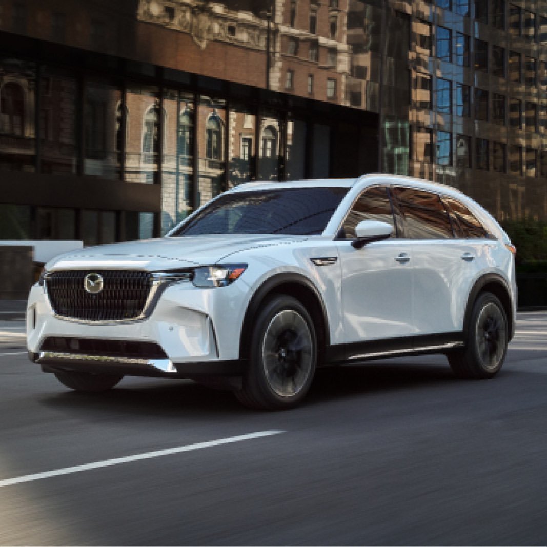 A white Mazda CX-90 SUV drives on an asphalt road in an urban area with buildings and trees on a sunny day. The vehicle is modern.