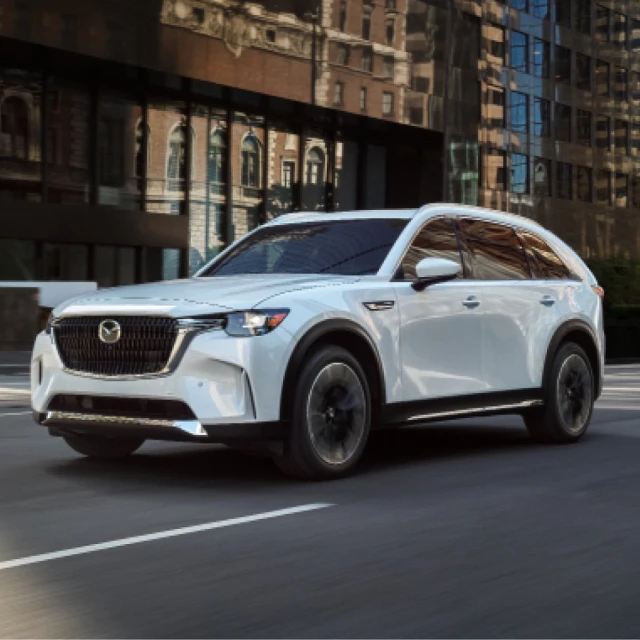 A white Mazda CX-90 SUV drives on an asphalt road in an urban area with buildings and trees on a sunny day. The vehicle is modern.