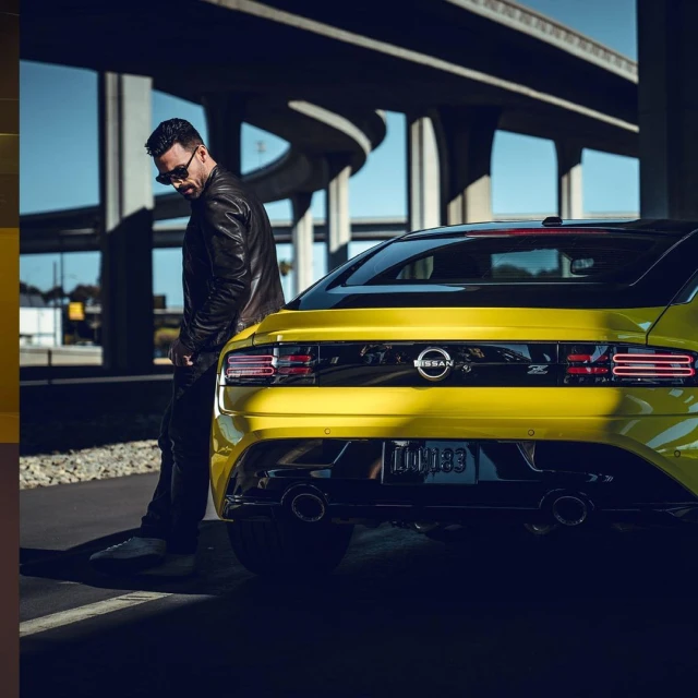 Man in black leather jacket leans on a yellow Nissan Z car under a concrete overpass on a sunny day with blue skies.