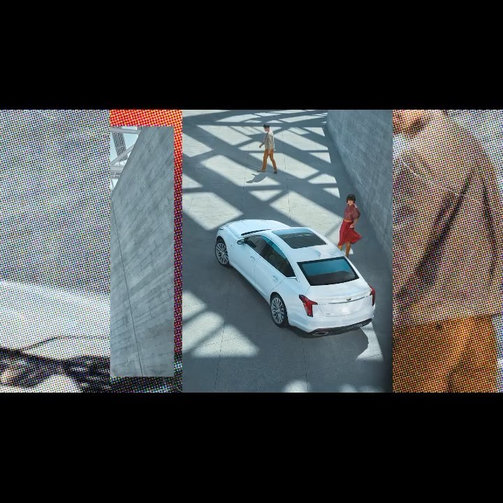High-angle shot of a white Cadillac CT5 parked in an urban environment, with two people walking nearby. The car and environment are modern.