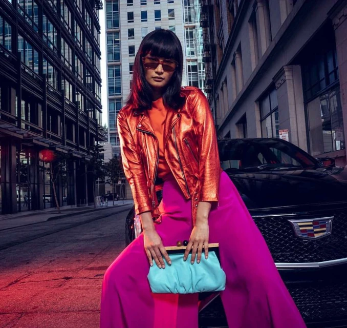 Woman in metallic jacket and pink pants poses on a city street near a Cadillac, holding a blue clutch with buildings in background.