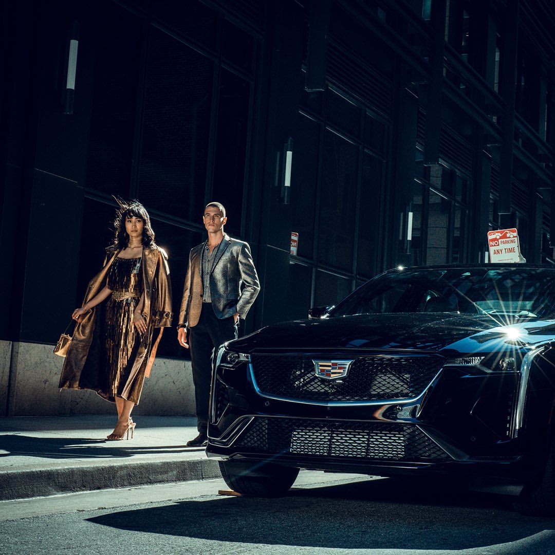 A black Cadillac car showcased alongside a man and a woman in evening wear on a city street in the evening with dark colors.
