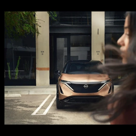 Bronze Nissan Ariya electric SUV parked in front of a modern building with architectural details, street level shot with partial view of person.