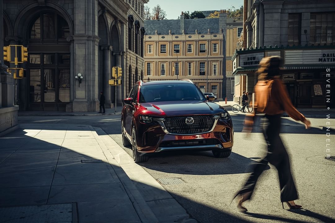 Burgundy Mazda CX-90 SUV parked on city street with pedestrian crossing the road on a sunny day, featuring architecture and traffic lights.