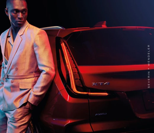 An adult male poses in a light-colored suit next to a red Cadillac XT4 car against a dark backdrop in a studio shot.