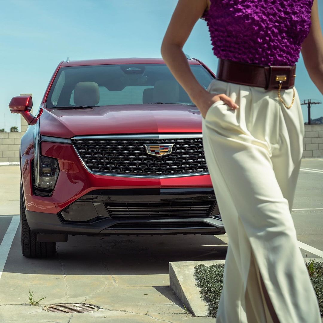 Red Cadillac SUV parked in an outdoor parking lot with an adult female partially in frame with cream pants and a purple top.