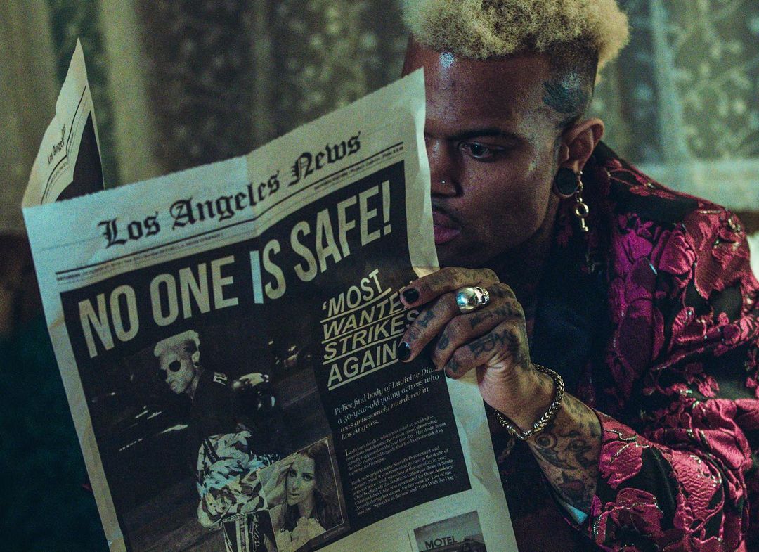 Tattooed adult male with bleached hair reads Los Angeles News newspaper headline "NO ONE IS SAFE!" with photos, wearing patterned jacket.