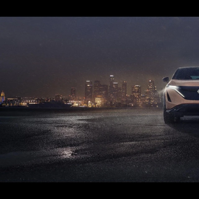 Bronze Nissan Ariya electric SUV parked in an urban setting against a backdrop of a lit-up city skyline at night.