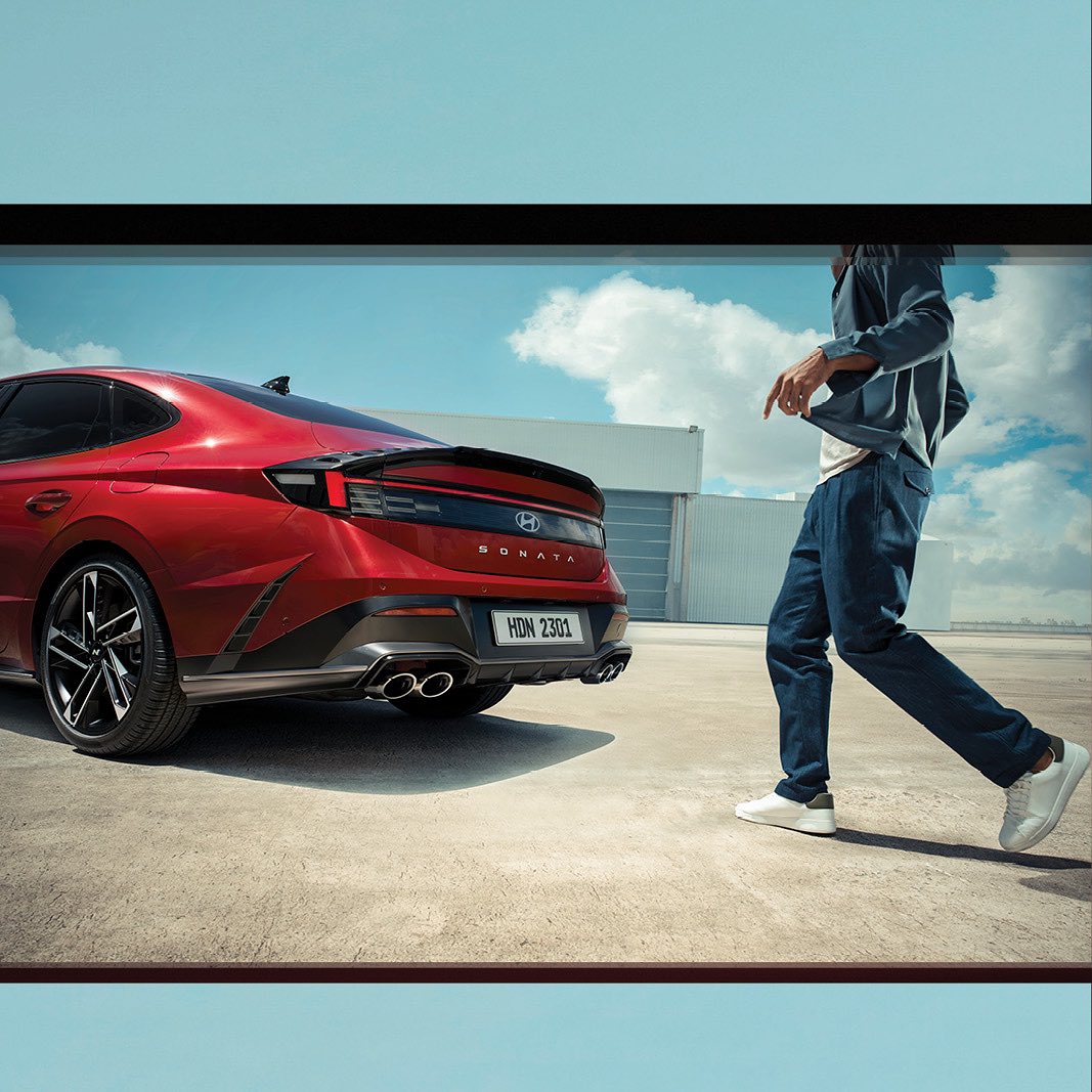 Red Hyundai Sonata sedan parked next to an adult male walking on a concrete lot under a partly cloudy sky, automobile advertisement.