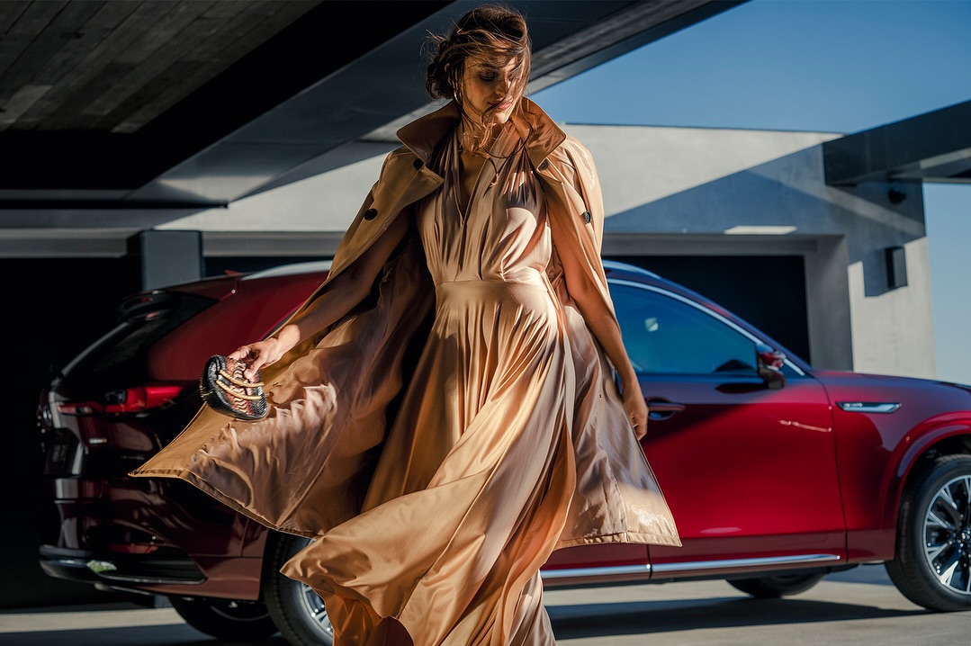 Brunette model in long tan coat and dress stands near a parked red SUV in front of a modern home with a concrete facade.