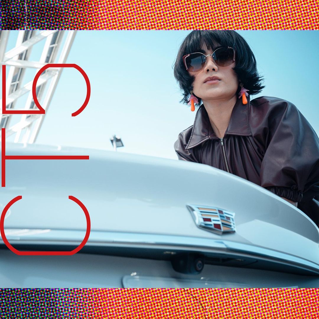 A woman in sunglasses leans on a white car with a modern art-inspired graphic overlaying the scene. The sky is visible in the background.