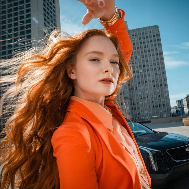 Fashionable woman with red hair poses next to a black car with city buildings in the background, showcasing modern style and urban environment.