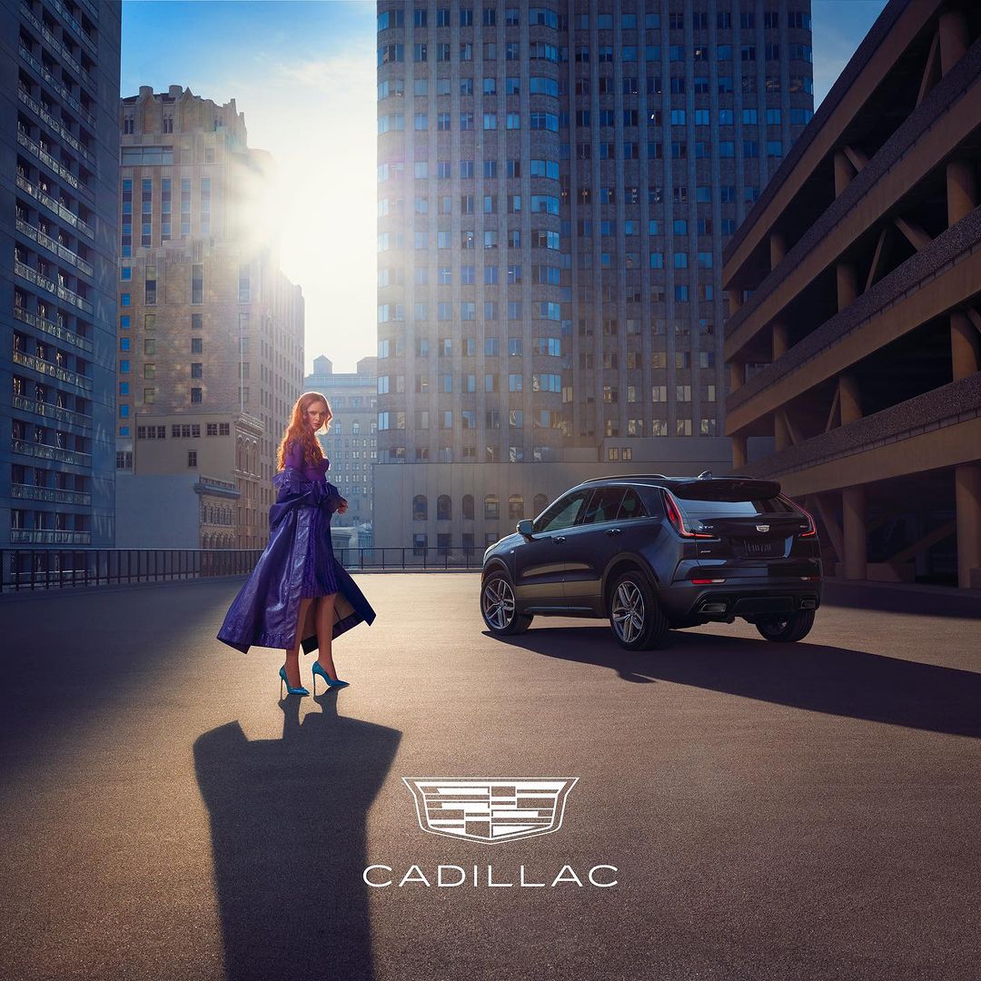 A woman stands near a black Cadillac SUV against an urban backdrop with skyscrapers, showcasing fashion and automotive elements.