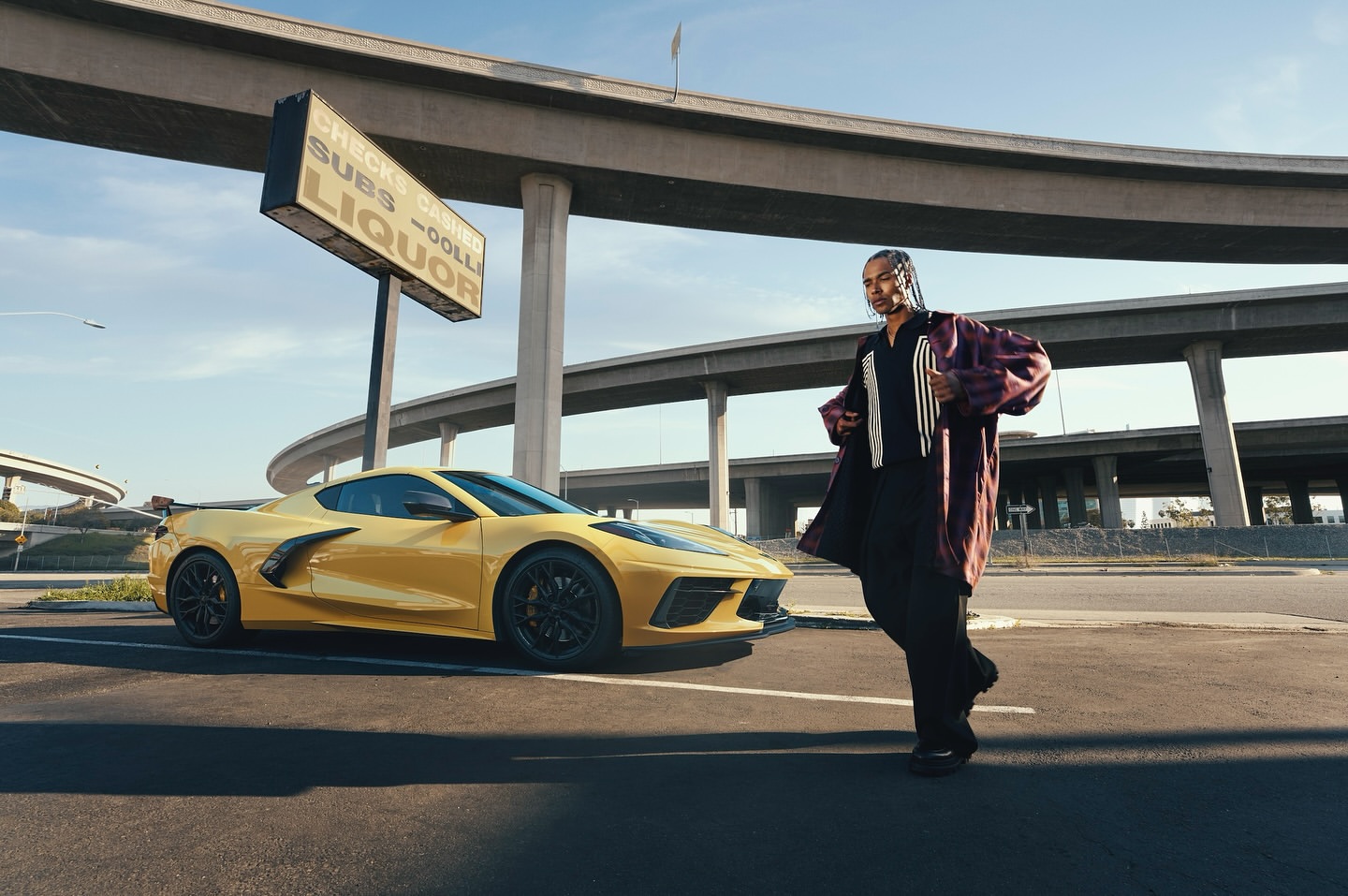 A yellow sports car is parked on an empty street with a man in a burgundy coat standing next to it during the daytime.