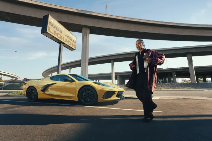 A yellow sports car is parked on an empty street with a man in a burgundy coat standing next to it during the daytime.