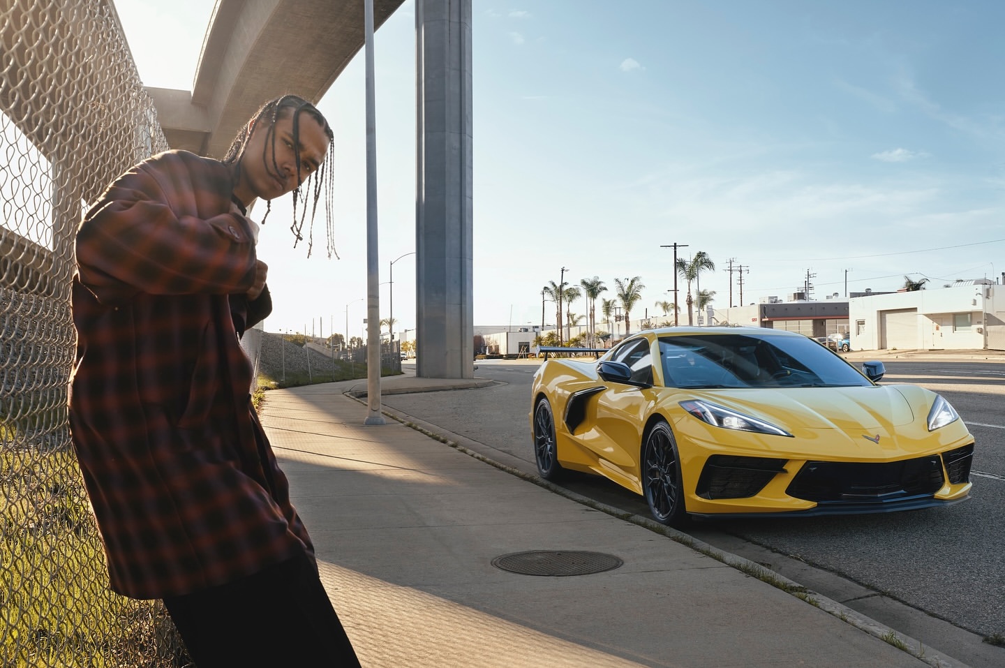 Adult male in red plaid shirt posing next to a yellow sports car in an urban environment with palm trees under a bright sky.