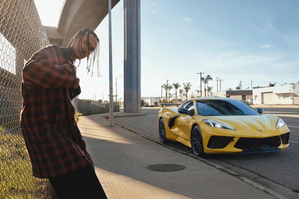 Adult male in red plaid shirt posing next to a yellow sports car in an urban environment with palm trees under a bright sky.