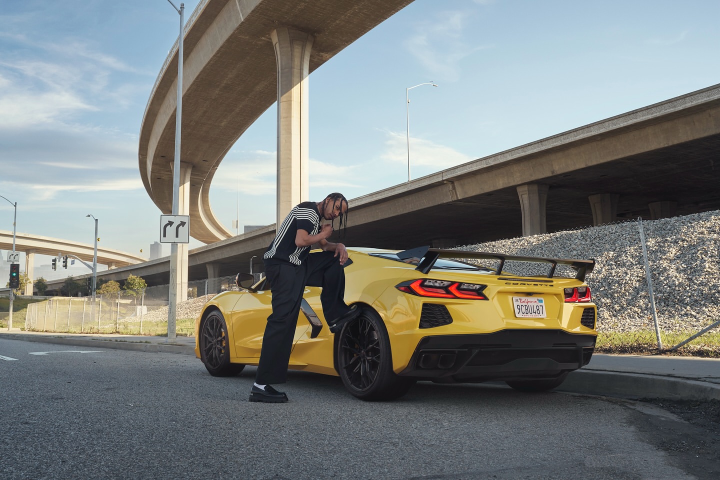 A man poses on a yellow Corvette Stingray against a modern urban backdrop featuring elevated highways under a sunny sky.