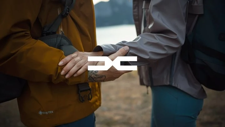 Close-up of two people holding hands outdoors with mountain backdrop. They wear jackets and backpacks, and a logo is centered.