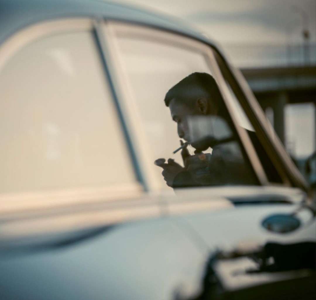 A man sits inside a vintage car, lighting a cigarette, seen through the car window in soft lighting with blurred background elements.