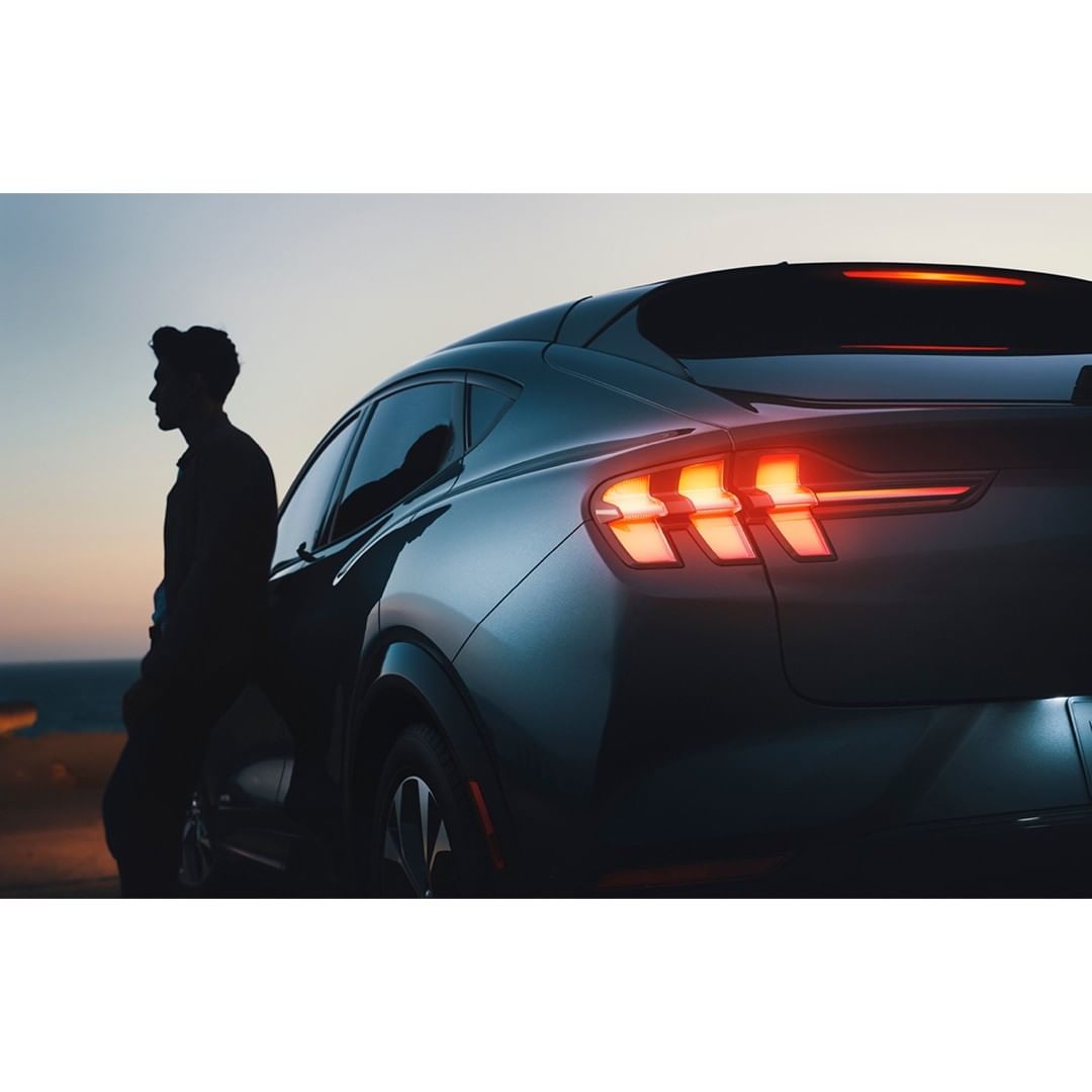 Sleek black electric car with illuminated taillights parked seaside at dusk, silhouette of a man standing nearby observing the view.