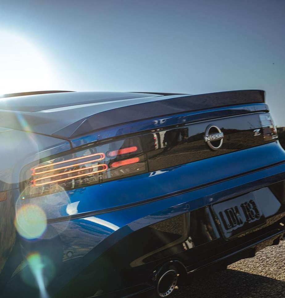 Rear view of a blue Nissan Z sports car on an outdoor paved surface, showcasing its taillights and logo under a clear sky.