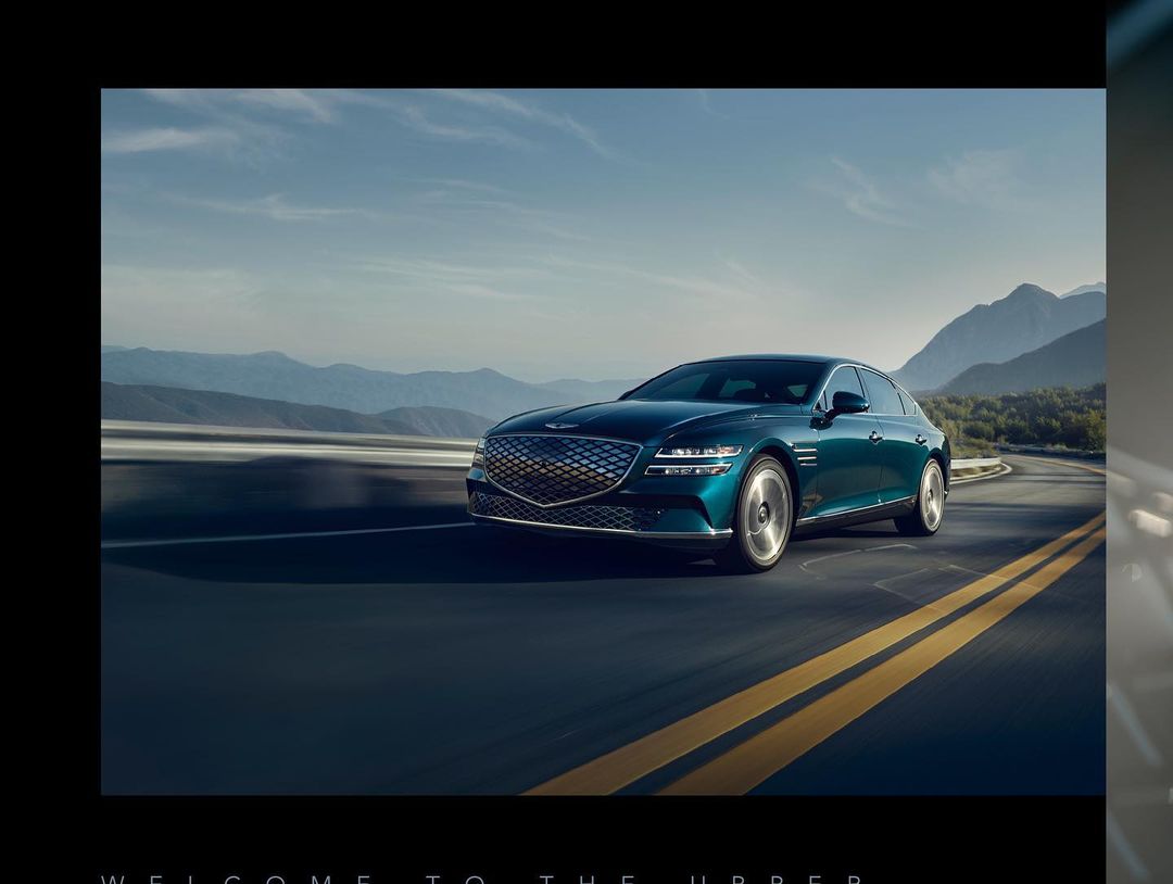 Turquoise Genesis sedan drives on winding asphalt road with yellow lines against a backdrop of mountains and blue sky.