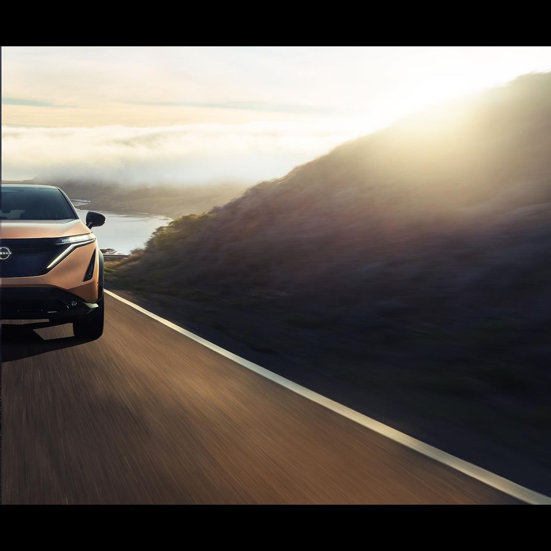 Bronze Nissan Ariya electric vehicle driving on a rural highway with mountains and a cloudy sky in the background in a sunny, outdoors scene.