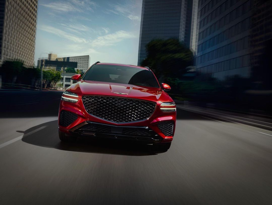 Dynamic low-angle shot of a red Genesis GV70 SUV driving on a city street with modern architecture, blue sky, and motion blur.