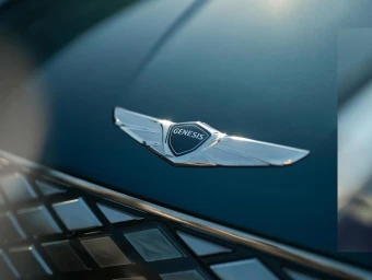 Close-up of a silver Genesis car badge and diamond-patterned chrome grille on a dark teal hood.