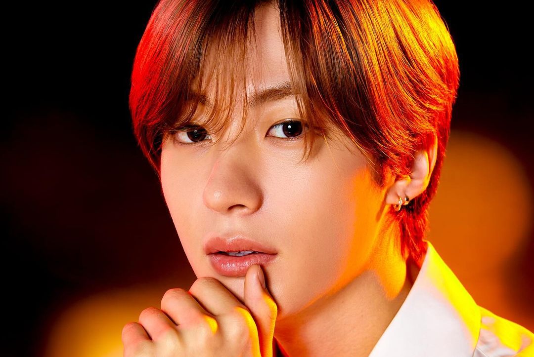 Close-up studio portrait of a young Asian man with red-brown hair, fair skin, light makeup, and hand near his face, set against a dark backdrop.