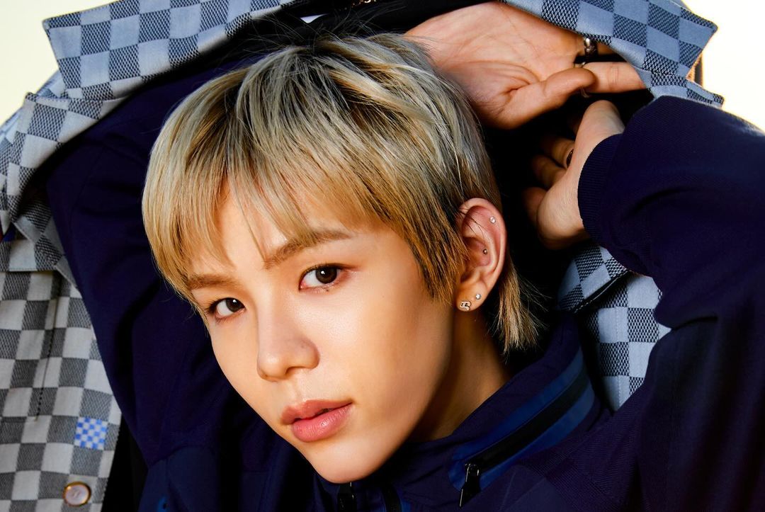 Close-up portrait of a young adult with blonde hair and brown eyes, wearing a dark blue jacket and patterned top, captured indoors.