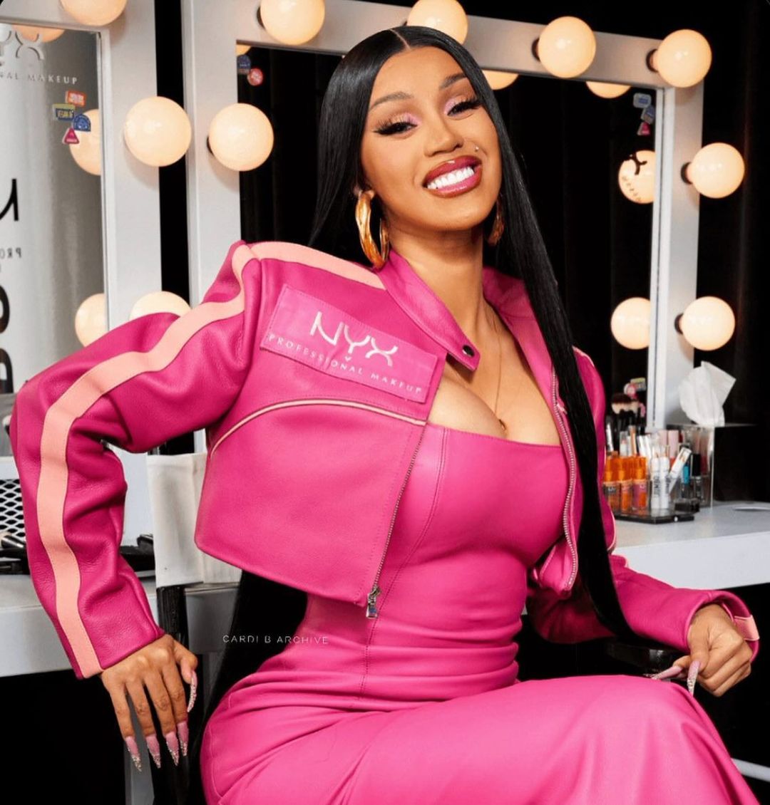 Close-up of an adult female celebrity, Cardi B, posing in a photo studio with a pink leather jacket, gold hoop earrings, and matching outfit.