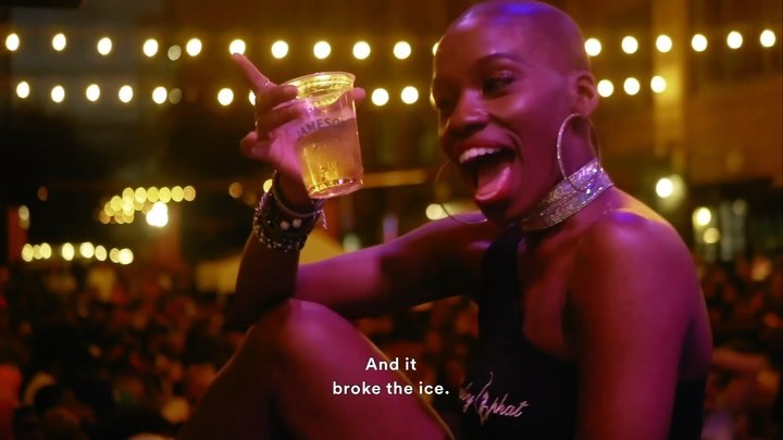 Woman holding a drink smiles at an outdoor party, surrounded by lights, with an excited facial expression and jewelry accents.