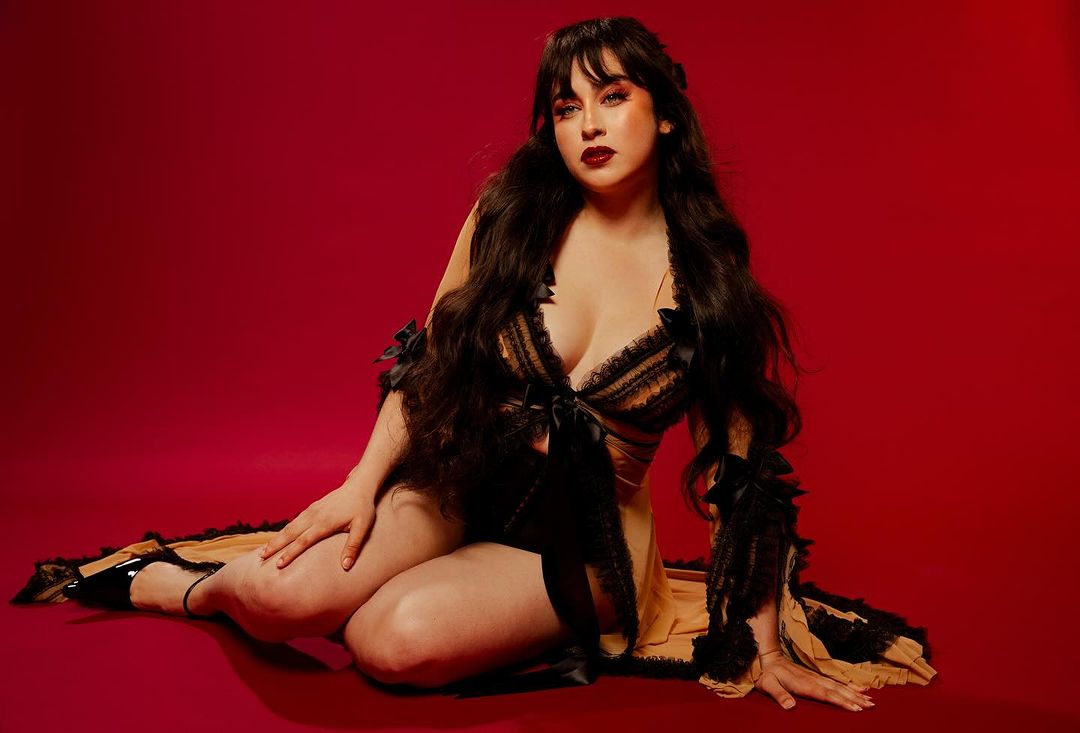 Brunette woman with dark lipstick and long hair poses in lingerie with black trim on a red background, seated with one leg outstretched.