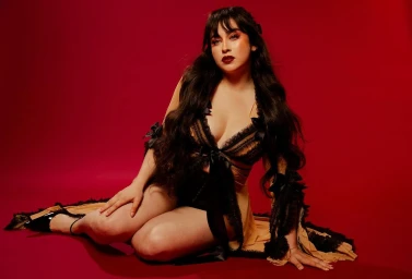 Woman with long dark hair wearing a beige and black lace outfit seated on a red studio backdrop