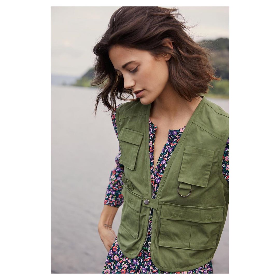 A woman with short brown hair wears a green vest over a floral dress outdoors near a body of water on an overcast day.