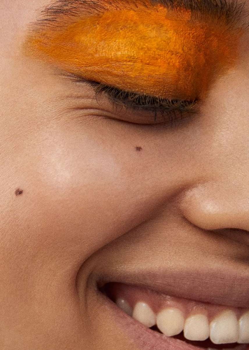Close-up portrait of a smiling adult female with orange eyeshadow. Eye is partially closed, revealing eyelashes. The skin is fair.