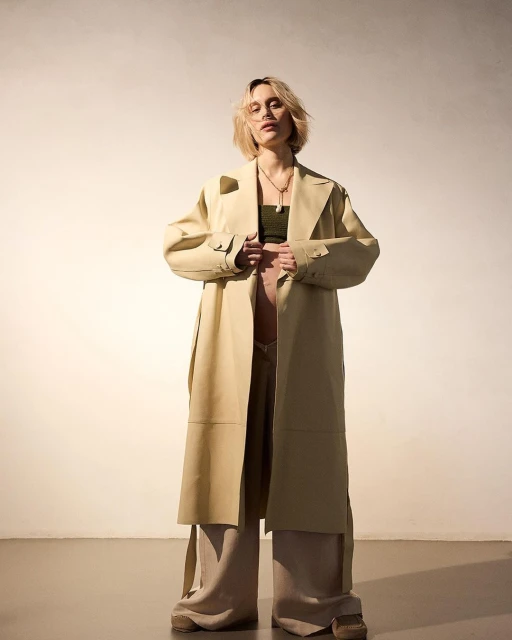 Stylish woman in a tan trench coat and wide-leg pants stands in a studio setting, showcasing a blend of modern fashion and timeless elegance.