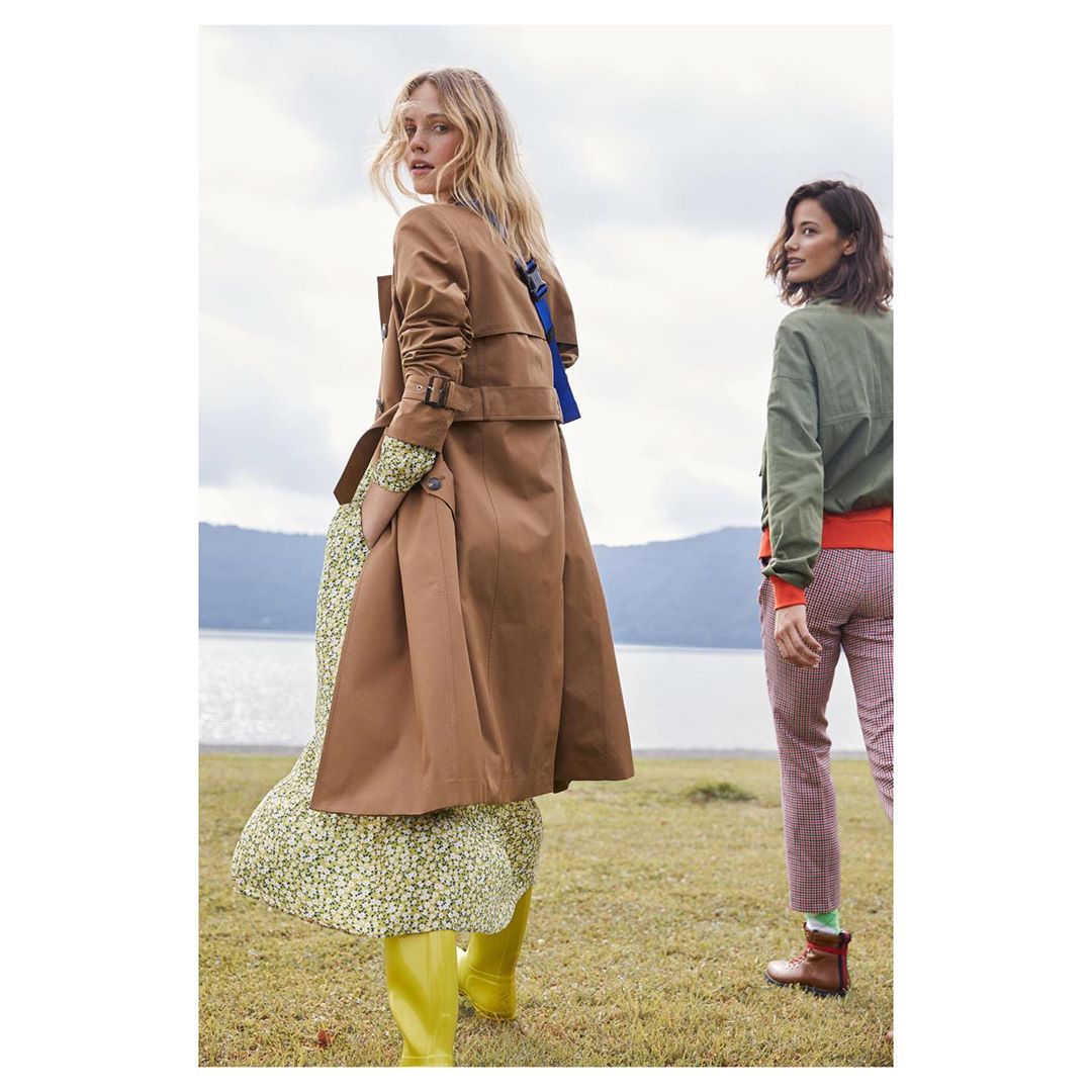 Two women in trench coat and olive bomber jacket walk in grass field with lake in background. Fashion outdoor editorial shoot.