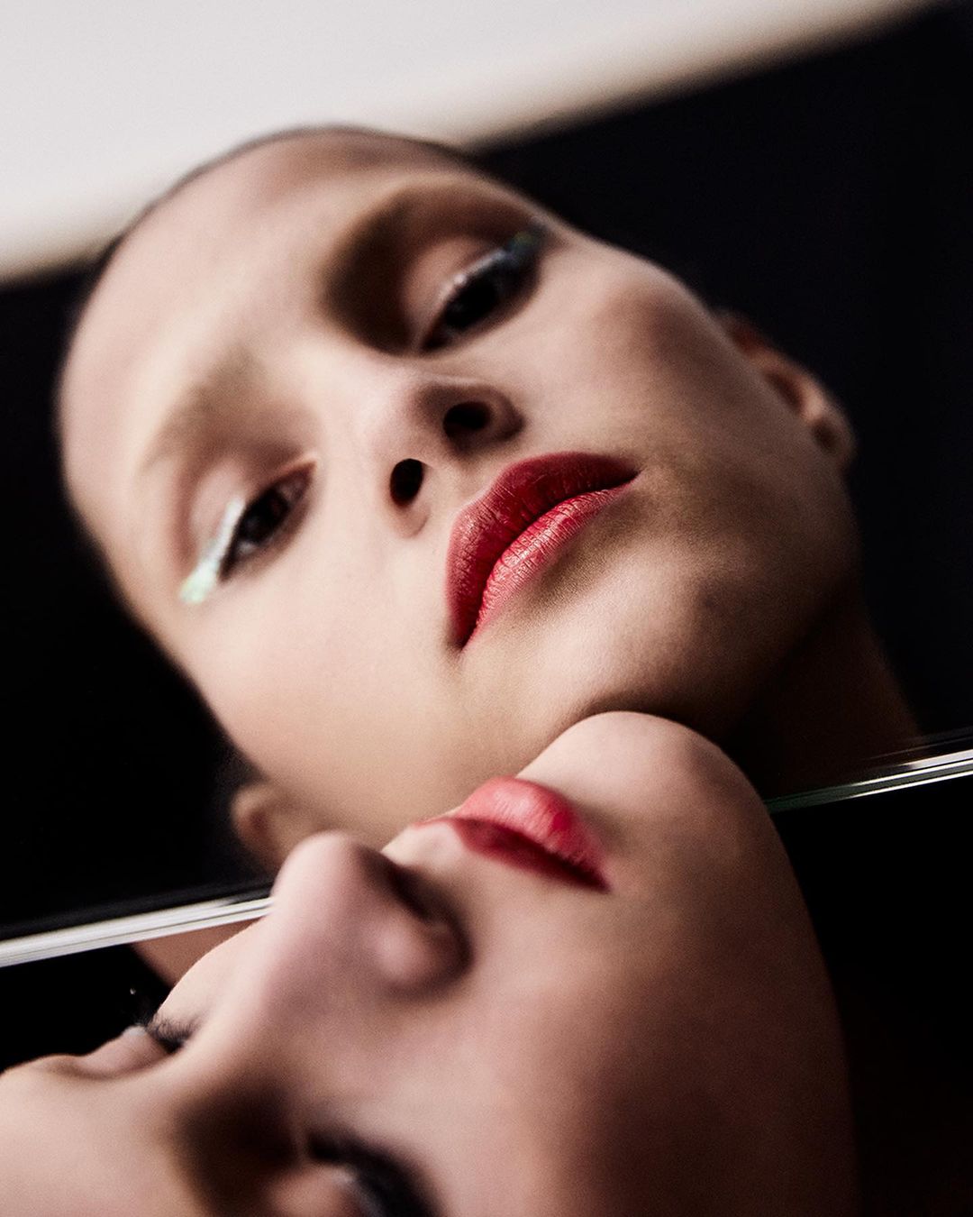 Close-up studio shot of a young fair-skinned woman with red lipstick and bright green eyeliner, reflected in a glass or mirror surface.