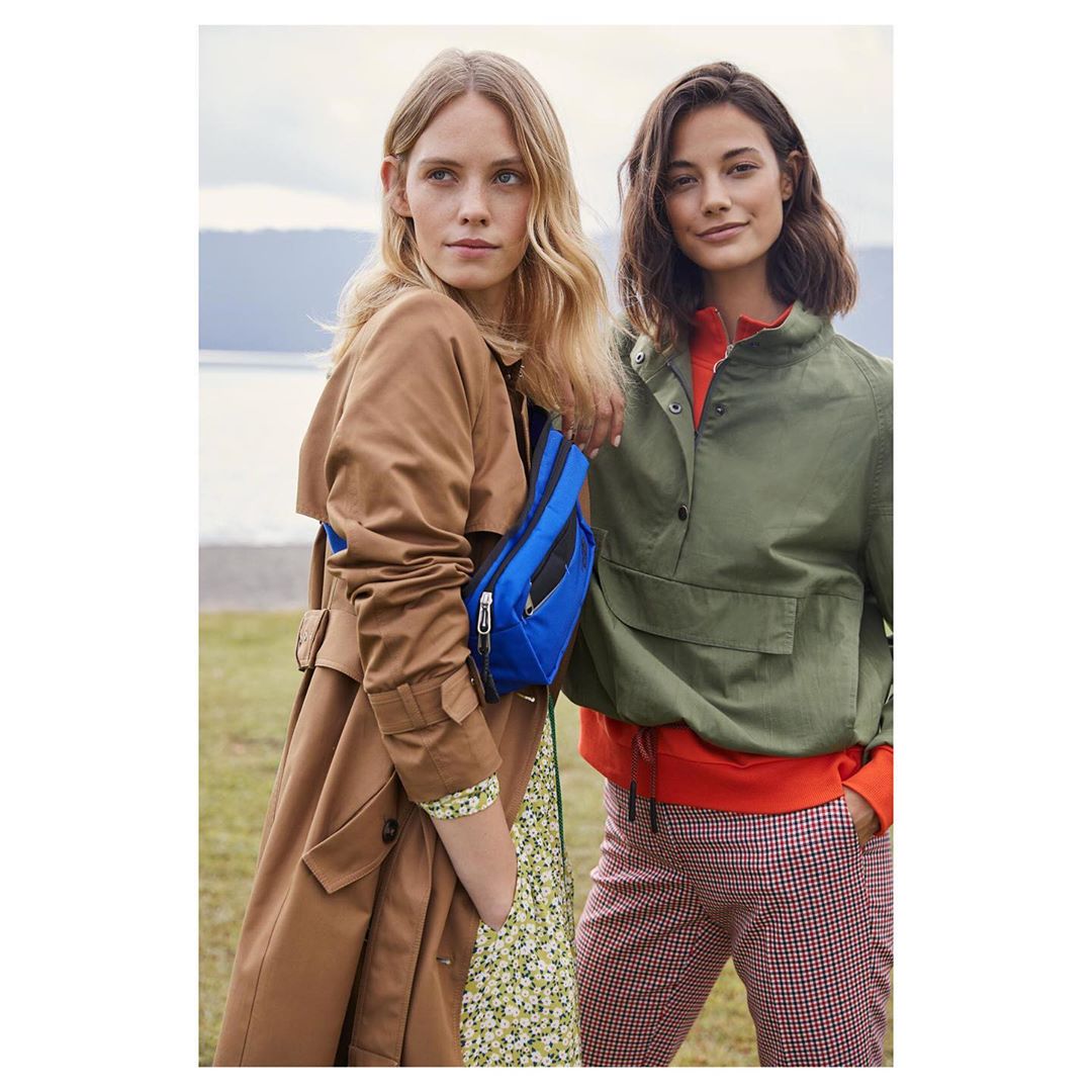Two women in fall fashion stand outdoors. One wears a trench coat and the other a green anorak, with a lake and hills in the background.